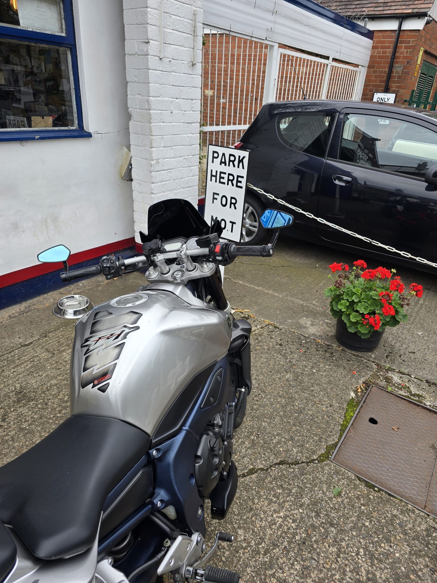 A silver and blue motorbike parked by the garage with red flowers and a Park here for MOT sign